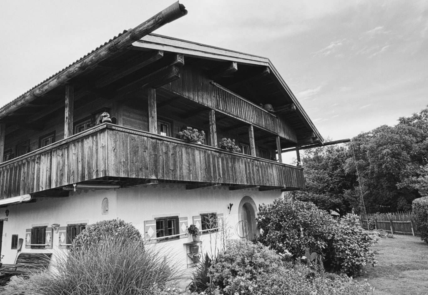 Traditionelles Holzhaus mit Balkon, umgeben von Pflanzen und Bäumen. Schwarz-weiß Aufnahme.